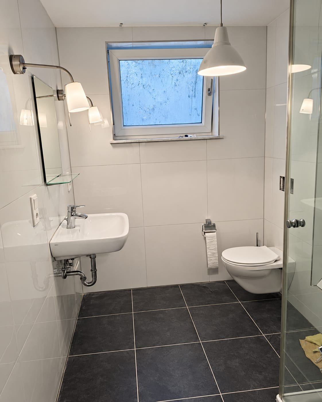 Dasselbe Bad fertig: große weiße Wandfliesen, dunkle Bodenfliesen, Waschtisch mit Spiegel und zwei Leuchten, wandhängendes WC, rechts die Dusche mit Glastür
