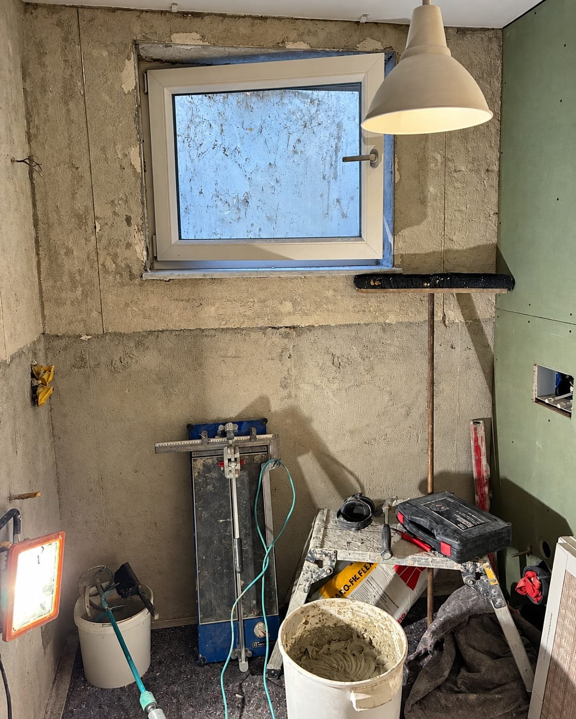 Bad im Rohbau vor den Fliesenarbeiten: verputzte Wände, Fliesenschneider, Eimer und Werkzeug, Fenster in der Rückwand