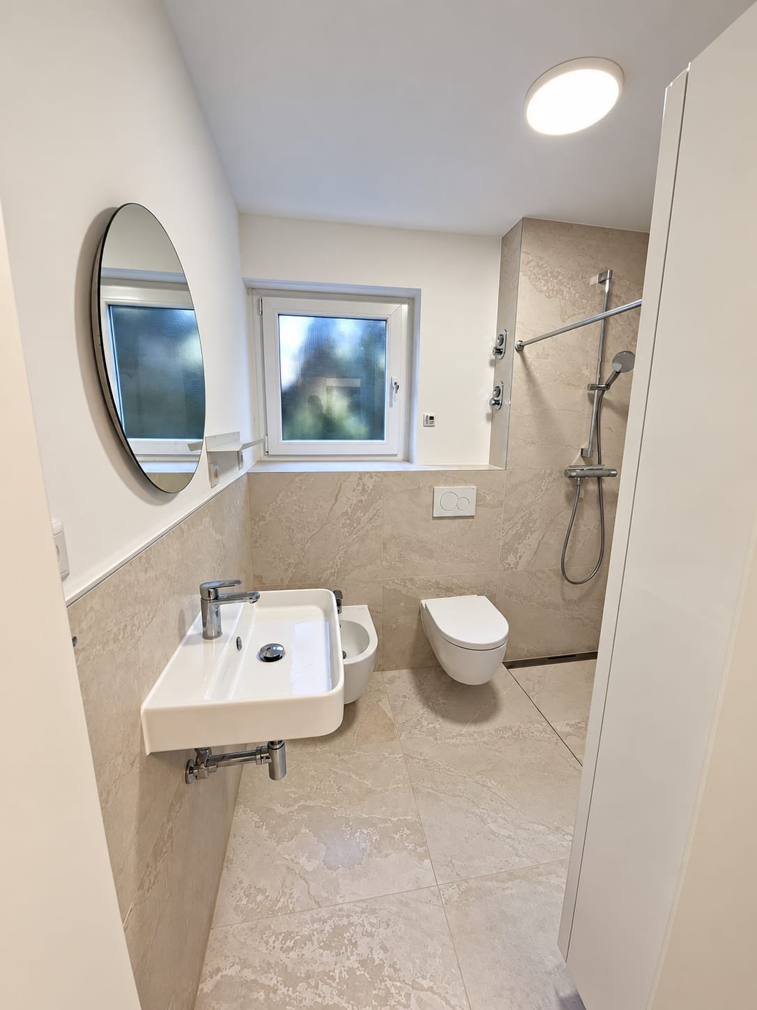 Dasselbe Bad fertig: Fliesen in beiger Steinoptik, bodengleiche Dusche mit Rinne, wandhängendes WC und Bidet, runder Spiegel über dem Waschtisch